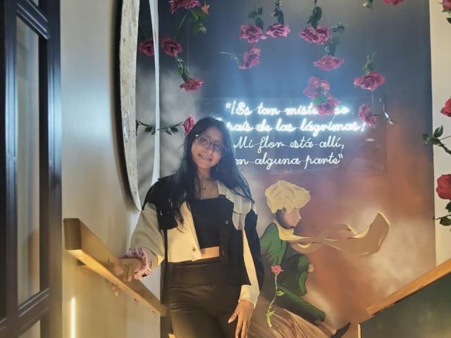 VioletXBloom with long black hair, glasses, white and black top, black leather pants, standing by a neon sign, smiling, hand on wooden railing, clock on wall.