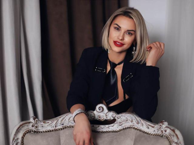 BelleLisaG with short blonde hair, red lipstick, black blazer, and black tie, leaning on an ornate white chair, right hand near her face, left arm on the chair.