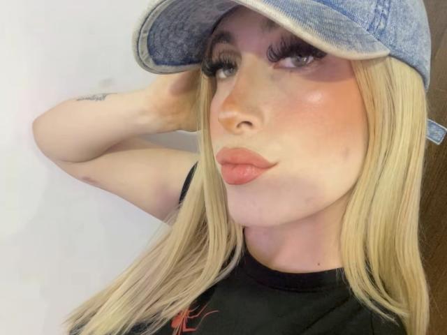 SammyViolet with blonde hair, wearing a blue denim cap, black shirt, and pouty pink lips, has a tattoo on her right arm and long eyelashes.