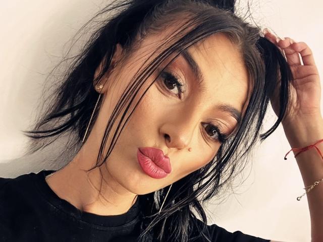 KinkyAmelie with pouty pink lips, brown eyes, and black hair in a messy ponytail, wearing a black shirt and red bracelet, hand in hair.