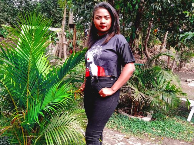 LinahSexy69 with dark hair, red lipstick, black t-shirt, red and white graphic, black jeans, hands in pockets, standing next to green palm leaves, forest background.