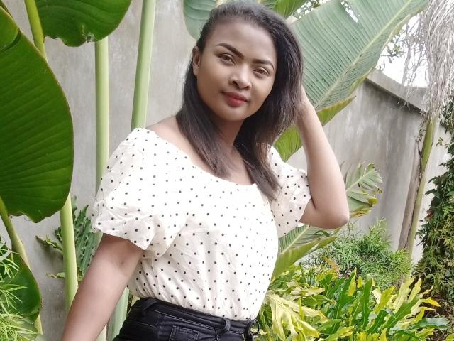 LinahSexy69 with dark hair, wearing a white polka dot top and black pants, standing in a green garden, hand in hair, smiling, showing small tits, cute ass.
