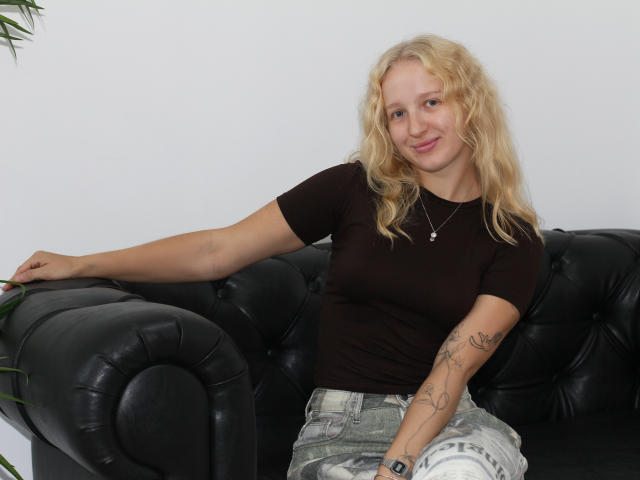 SummerBirch with blonde curly hair, brown t-shirt, white and grey jeans, tattooed arm, black leather couch, smiling, right arm on couch, left arm resting, pendant necklace.