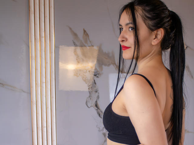 AriaBonnet with long black hair in a ponytail, wearing a black sports bra, showing her tits, red lipstick, and looking over her shoulder, standing near vertical blinds.