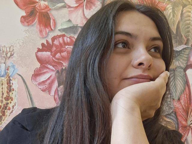 KatherinLatin with long dark hair, nose ring, and black shirt, resting her chin on her hand, smiling subtly, brown eyes, and smooth skin. Floral patterned wallpaper in background.