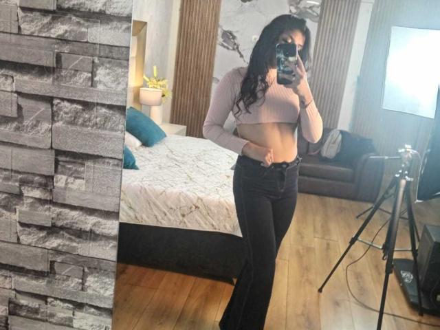 TatianaMorelli with long black hair, pink ribbed crop top, black high-waisted jeans, holding phone, taking mirror selfie, showing flat stomach, small tits, in modern bedroom.