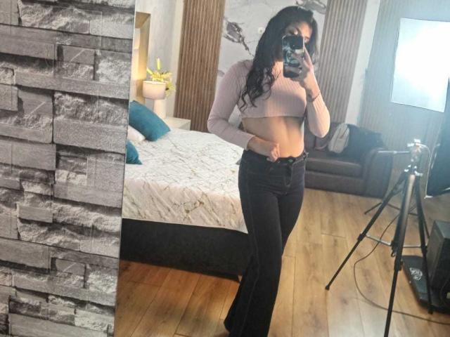 TatianaMorelli with long black hair, pink crop top, showing her flat abs, black flared jeans, taking a mirror selfie, holding a phone, in a wooden-floored room.