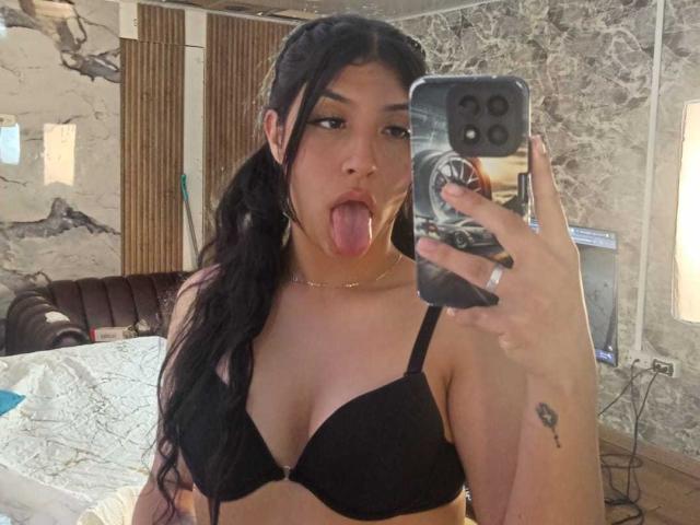 TatianaMorelli with long black hair, tongue out, wearing a black bra, taking a mirror selfie, showing her tits, holding a phone with a ring design, has a tattoo.