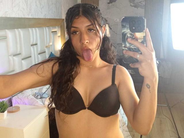 TatianaMorelli with long dark hair, sticking out her tongue, wearing a black bra, holding a phone, and a small tattoo on her right arm. Her tits are visible.