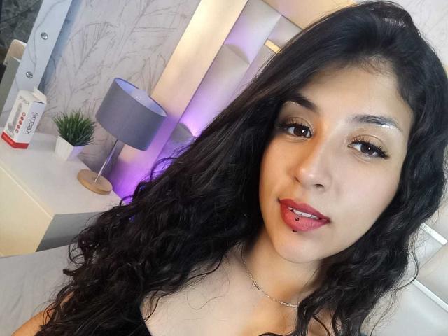 TatianaMorelli has long, wavy black hair, brown eyes, and is sticking her tongue out with a small metal ball piercing. She's wearing a black top and a silver necklace.