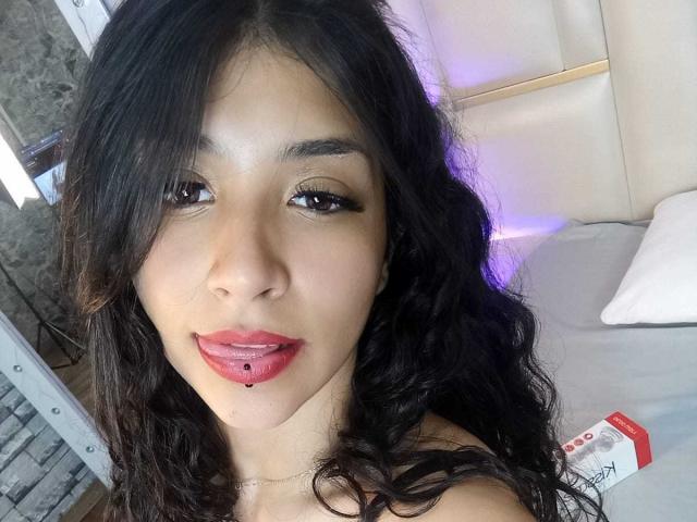 TatianaMorelli with long black wavy hair, light brown skin, dark eyes, red lipstick, and a small black lip piercing, sitting on a bed with white pillows.