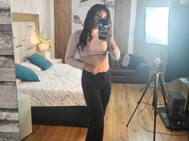 TatianaMorelli with long black hair, pink crop top, exposing her flat midriff, and black jeans, taking a mirror selfie in a bedroom with a bed and tripod.