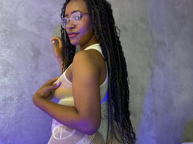 NaomiReyes with long braids, wearing clear glasses and a white fishnet top, showing her tits, standing side-on, hand on chest, looking at camera.