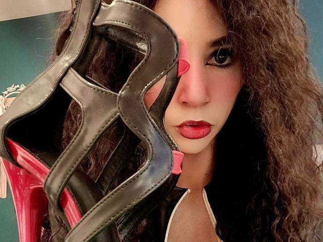 SexyKonnyX, avec ses cheveux bruns bouclés, son rouge à lèvres rouge et son masque noir dissimulant partiellement son visage, tient un masque noir et rose devant elle. Elle a un regard séduisant..