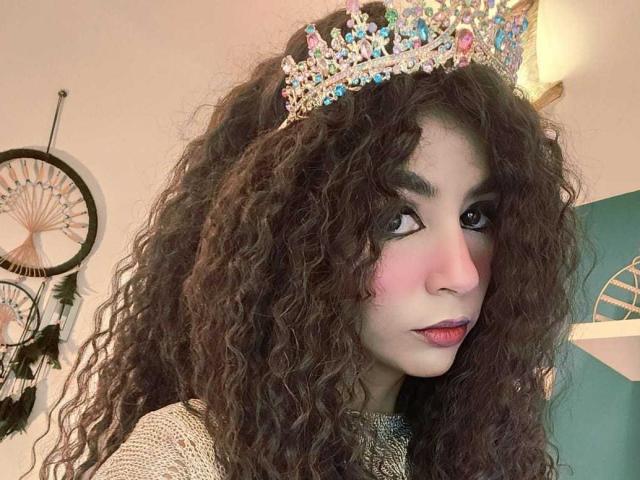 SexyKonnyX, avec ses cheveux bruns bouclés, ses joues roses et sa couronne scintillante et colorée, porte un pull gris, a du rouge à lèvres rose et des yeux foncés, et regarde sur le côté..