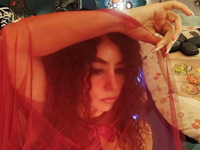 SexyKonnyX avec des cheveux bruns bouclés, portant une robe rouge transparente, levant un bras avec de longs ongles blancs, une lumière bleue dans les cheveux, des lèvres roses, regardant de manière séduisante l'appareil photo..