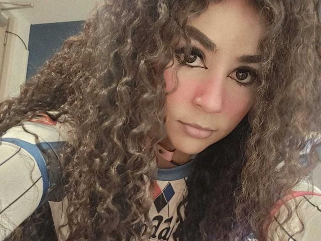 SexyKonnyX with curly brown hair, pink blush, and brown eyes, wearing a striped shirt, looking at the camera with a slight smile. Her nose is small and her lips are full.