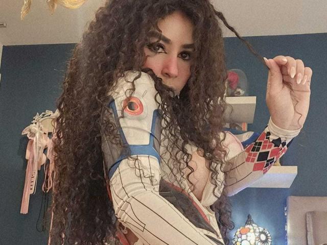SexyKonnyX avec de longs cheveux bruns bouclés, portant une tenue futuriste blanche et bleue, tenant un fil noir, avec un nez rose, un mur bleu en arrière-plan, et un ruban rose sur la gauche..
