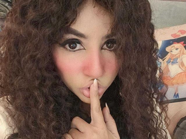 SexyKonnyX avec des cheveux bruns bouclés, un blush rose et un eye-liner foncé, suçant son doigt avec un doigt dans la bouche, regardant directement l'appareil photo..