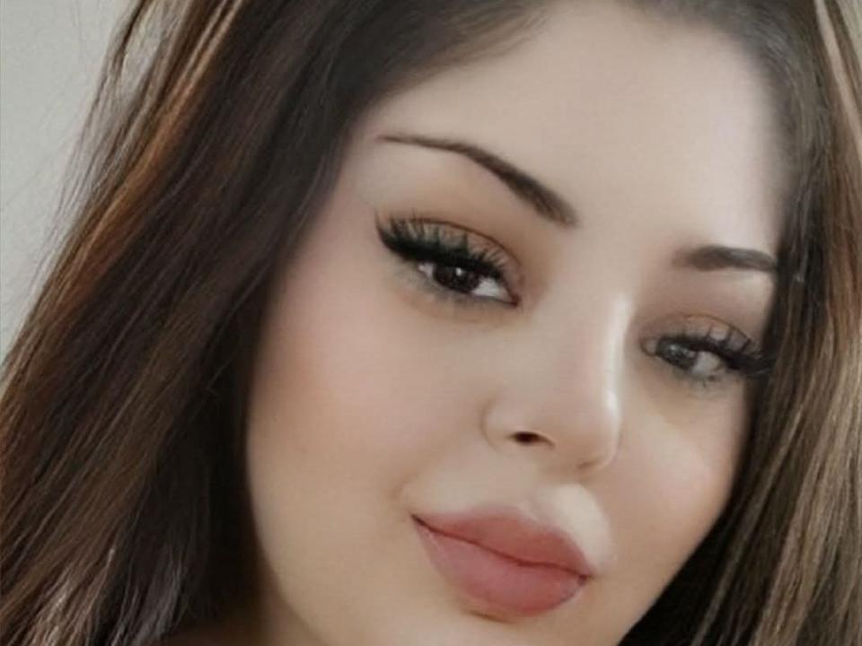 AlinaSweetX has light skin, brown eyes with long lashes, and full pink lips. She has dark brown hair and a slight smile. Her eyebrows are thick and well-groomed.