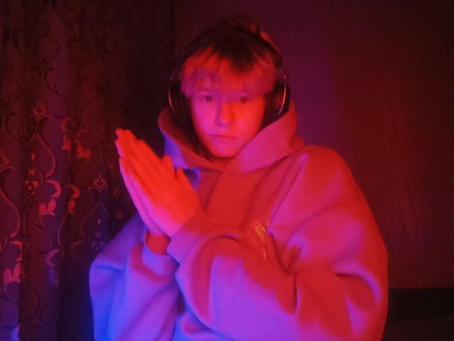 JimmieHot with pink and purple-tinged skin, wearing a fluffy pink hoodie and headphones, claps hands together, looking slightly confused or surprised, in dim, colorful lighting.