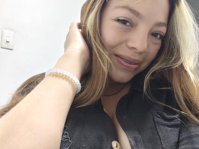 ScarlettAmelia with blonde hair, black shirt, and white bracelet, touching her head, smiling with pierced lower lip, showing a bit of her tits. She's looking at the camera.