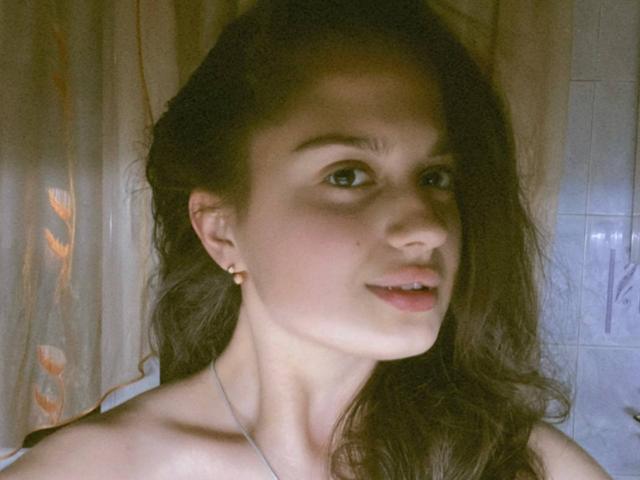 TaliaSeduce ha lunghi capelli castani, occhi verdi e un leggero sorriso, indossa piccoli orecchini e una collana, mostrando le spalle scoperte e la sua pelle delicata..