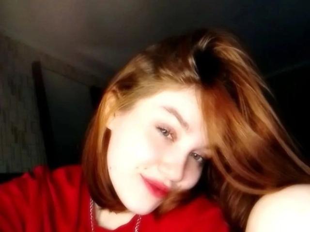 EllieFire has pale skin, red hair, and red lipstick, wearing a red shirt, smiling at the camera with a slight tilt to her head, in a dark room.
