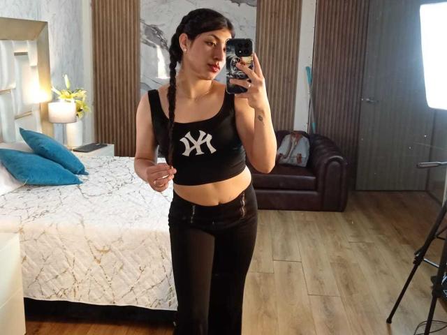 TatianaMorelli with braided hair, black sports bra showing small tits, and NYC logo, black pants, taking a mirror selfie in a bedroom with a bed and brown chair.