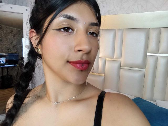 TatianaMorelli with dark hair in a braid, brown eyes, red lipstick, black bra, silver necklace, and facial piercings, smiling at the camera. Her tits are partially visible.