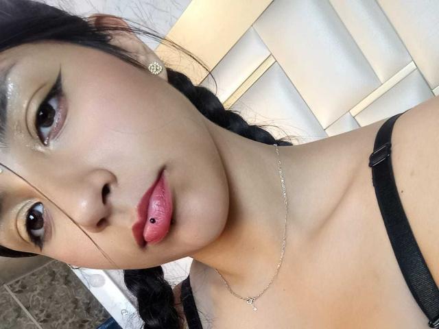 TatianaMorelli has glossy brown eyes, black braided hair, and pink lipstick with a lip piercing. She's wearing a black strap, and a delicate necklace. Her skin is light.