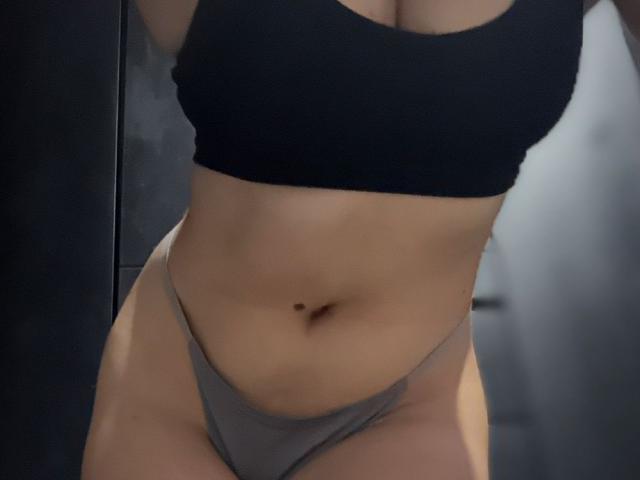 AnastasiaEvansX has large tits in a black sports bra and a grey thong, showing a tan belly with a small belly button. Her body is slightly curvy.