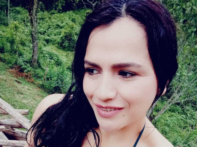 CameronViviane has dark wet hair, light skin, and is smiling with small tits visible. She's outdoors with greenery and a log in the background.