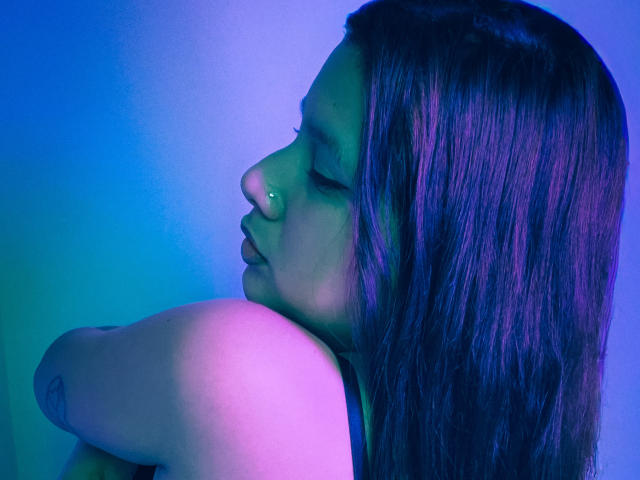 LadyStarOne con los ojos cerrados, un piercing en la nariz y un cabello largo de color púrpura-azul con reflejos, sosteniendo sus pechos, con un tatuaje visible en el brazo superior, sobre un fondo degradado azul..