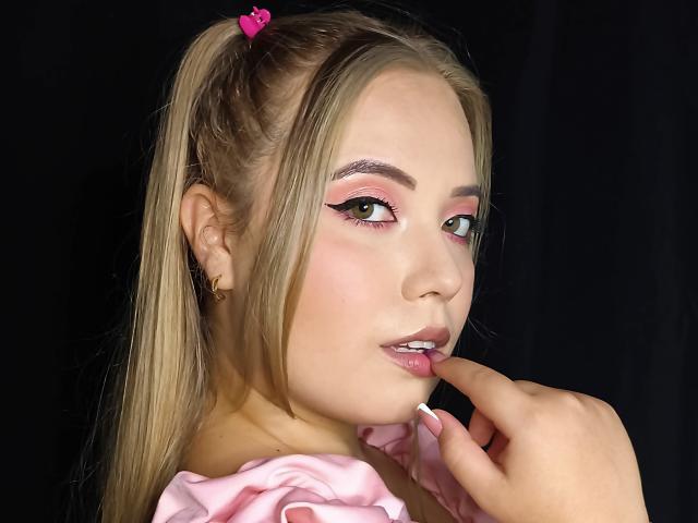 ZoeTobon69 with blonde ponytail, pink heart clip, heavy pink eyeshadow, glossy pink lips, finger near lip, pink ruffled top, black background, soft light, cute, sexy.