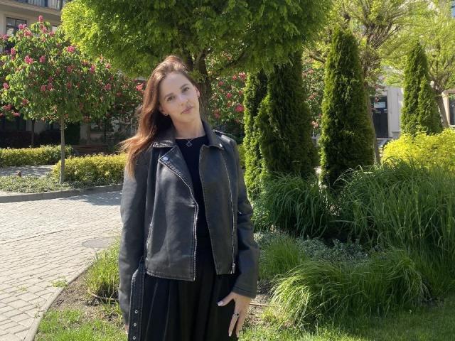 LoliLuv has long brown hair, wearing a black leather jacket over a black dress, standing in a sunny garden with green trees and pink roses. Her ass is not visible.