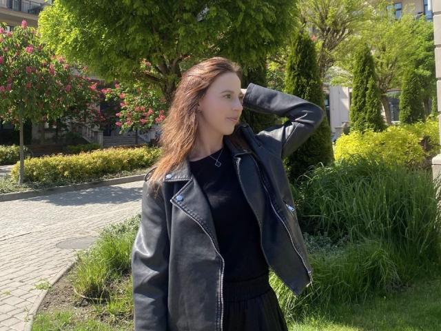 LoliLuv with brown hair, wearing a black leather jacket and dress, stands in a garden, hand in hair, looking to the side, sunny, greenery, grass, and trees around.