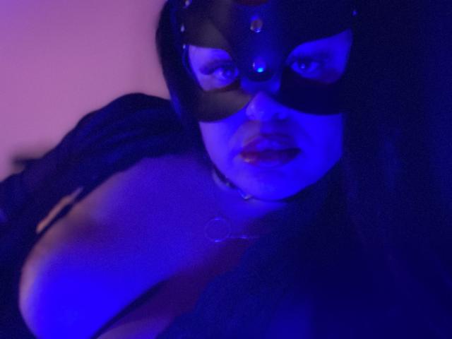 AlmaMilao in a black mask with blue lighting, showing her tits in a low-cut top, wearing a choker with a ring, and looking at the camera with parted lips.
