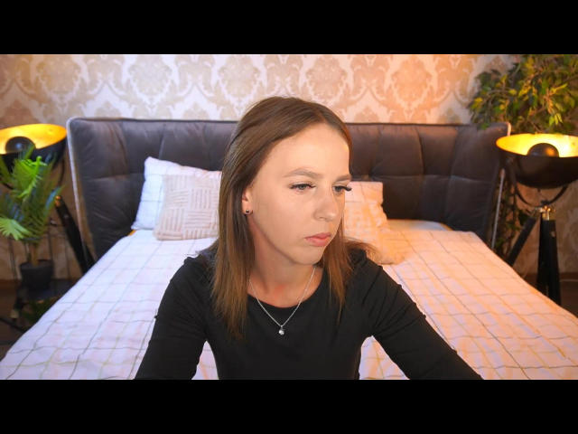 LoliLuv with shoulder-length brown hair, wearing a black shirt, necklace, and ear studs, sitting on a bed with a black headboard, white pillow, and yellow lamp on each side.