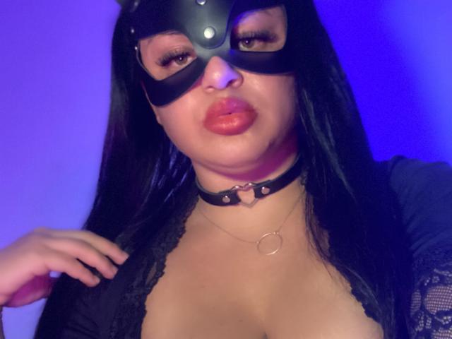 AlmaMilao with black leather mask, pouty orange lips, long black hair, black lace top, choker with ring, and hands near chest, under blue light. Boobs visible, sexy.