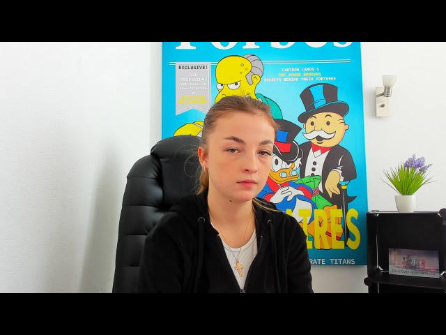 Aldovia, con i capelli biondi, un viso leggermente corrugato, indossa una felpa nera e una maglietta bianca, è seduta su una poltrona in pelle nera, con un poster dei "Simpsons" dietro di lei, sulla parete destra..
