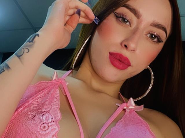 SarahLuxe, avec un soutien-gorge en dentelle rose, du rouge à lèvres rouge et des boucles d'oreilles pendantes, touchant sa paupière avec un applicateur à mascara violet, montrant un tatouage sur son bras droit..
