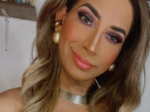 SashaSweetHot has purple eyeshadow, pink lipstick, and wavy blonde hair with pearl earrings and a silver necklace, smiling with a cute, sexy face.