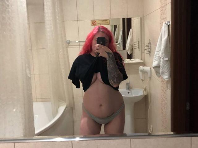 FortuneCarmen with pink hair, black shirt lifted, showing her tits, gray panties, tattooed arm, taking a mirror selfie in a bathroom.