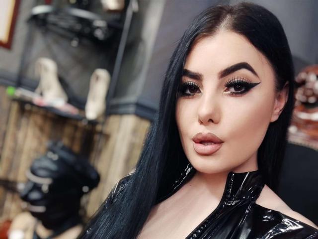 MelissaGlow has long black hair, dark eyebrows, heavy eyeliner, and glossy pink lips. She's wearing a shiny black latex top, looking serious with a slight pout.