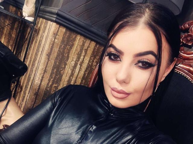 MelissaGlow with dark brown hair, thick eyebrows, long eyelashes, and pink lips, wearing a black leather zip-up, looking at the camera, sitting against a wooden wall.