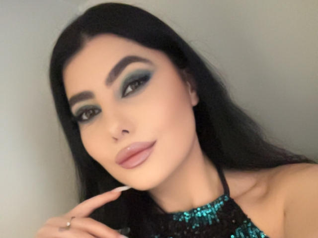MelissaGlow has black hair, green eyeshadow, and glossy pink lips. She's wearing a black sequin halter top, touching her chin with a ring on her finger.
