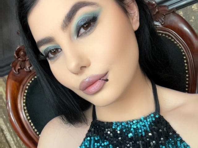 MelissaGlow has black hair, blue-green eyeshadow, brown eyes, a black sequin top, and a lip piercing. She's looking at the camera with a slight pout.