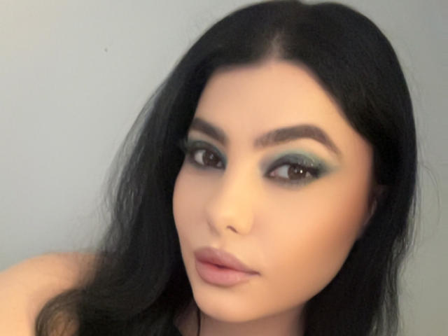 MelissaGlow has dark, wavy hair, green eyeshadow, and full pink lips. Her brown eyes are slightly upturned, and she has a subtle, sexy, confident expression.