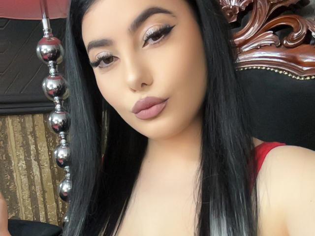 MelissaGlow has long black hair, brown eyes with winged eyeliner, full pink lips, and a red dress. Her ass is partially visible in the background.